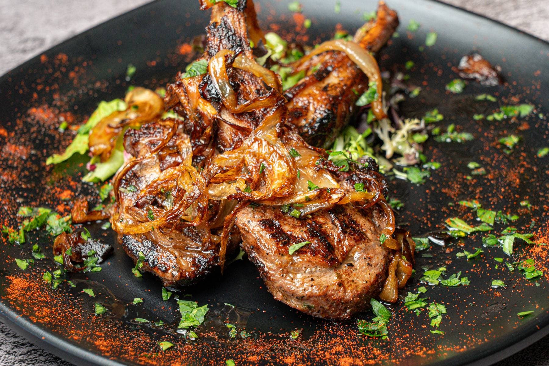 Grilled lamb chops blanketed in caramelized onions and parsley, plated black with sumac dust